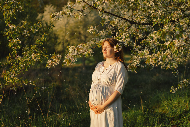 Ανοιξιάτικο πορτραίτο εγκύου. Μια όμορφη νεαρή έγκυος γυναίκα με λευκό φόρεμα περπατάει στον ανοιξιάτικο κήπο. Ευτυχισμένη εγκυμοσύνη. Άνοιξη - Φωτογραφία, εικόνα