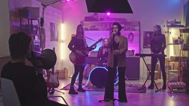 Slowmo of young all-female rock band recording music video in studio performing song staying on stage with neon lighting while their music producer giving commands in megaphone - Záběry, video