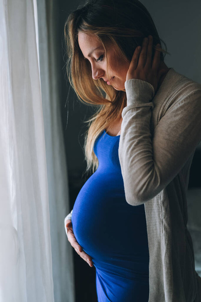 Giovane donna caucasica incinta posa in casa toccando pancia - Foto, immagini