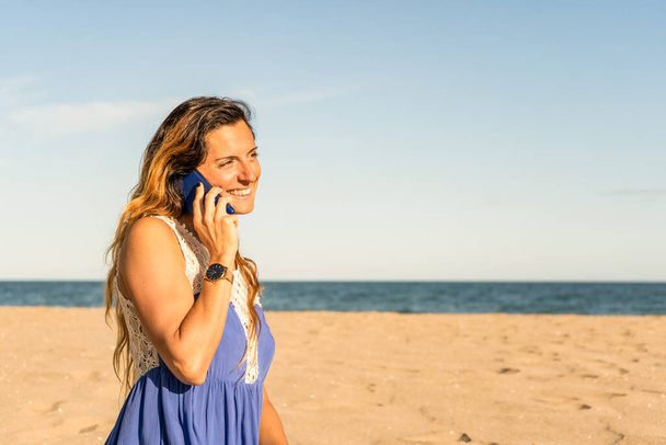Joyeuse femme élégante parlant sur un téléphone portable sur la plage au coucher du soleil.Femme utilisant un smartphone, passer un appel pendant les vacances d'été destination. - Photo, image