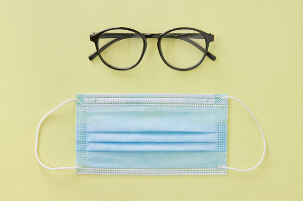 Top View Black Glasses and Blue Face Mask on Pastel Yellow Minimalist Background - Photo, Image