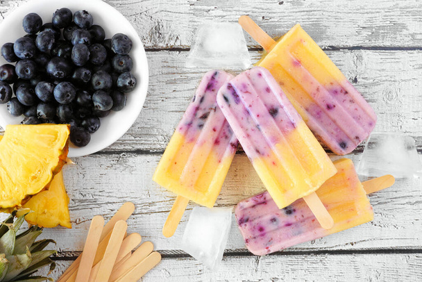 Pineapple and blueberry ice pops in a cluster. Above view table scene over a rustic white wood background. - Photo, Image