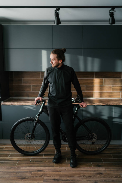A hipster man in a black tracksuit and sneakers is standing in the kitchen next to a black bike. Life style. Preparing for a bike ride. Weekend planning - 写真・画像