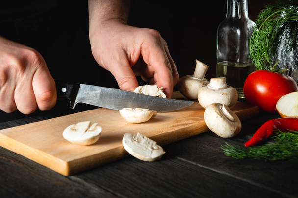 Szef kuchni kroi grzyby nożem, aby przygotować pyszne jedzenie. Zestaw warzyw na zabytkowym stole kuchennym w restauracji lub kawiarni. Pyszny przepis na śniadanie lub obiad - Zdjęcie, obraz