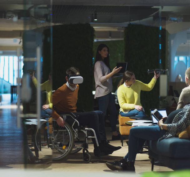 Disabled businessman in a wheelchair at work in modern open space coworking office on team meeting using virtual reality goggles and drone assistance simulation or brainstorming. Effective teamwork of - Zdjęcie, obraz