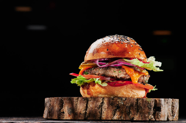 Rundburger met kaas, tomaten, rode uien, komkommer en sla op houten snit op zwarte achtergrond. Burger menu. Ruimte voor tekst. Ongezond voedsel. - Foto, afbeelding