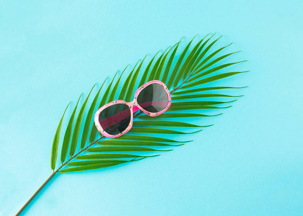 緑のヤシの葉の上にピンクのサングラス。ライトブルーを基調としたミニマルな夏のコンセプト - 写真・画像