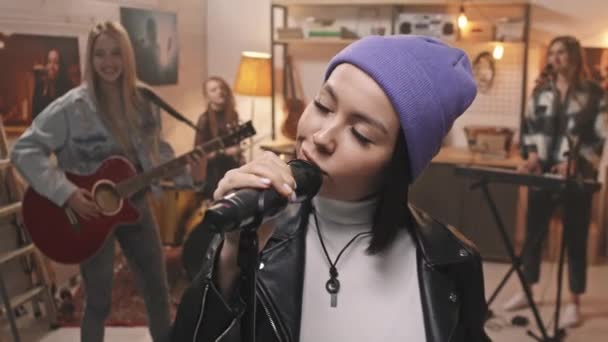 Slowmo detailní záběr cool rocková dívka v nóbl fialové čepice hledá sebevědomě na kameru při zpěvu v mikrofonu s kolegyněmi hraje na hudební nástroje v pozadí v útulném retro studiu - Záběry, video