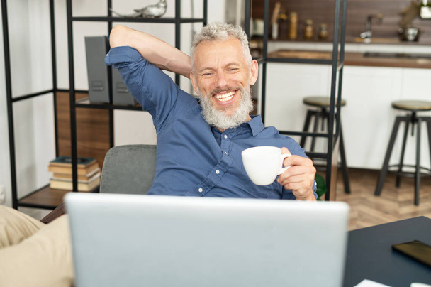 Cheerful mature male employee enjoys morning coffee at the workplace in the office - Valokuva, kuva