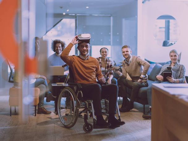 Disabled businessman in a wheelchair at work in modern open space coworking office on team meeting using virtual reality goggles and drone assistance simulation or brainstorming. Effective teamwork of - Фото, зображення