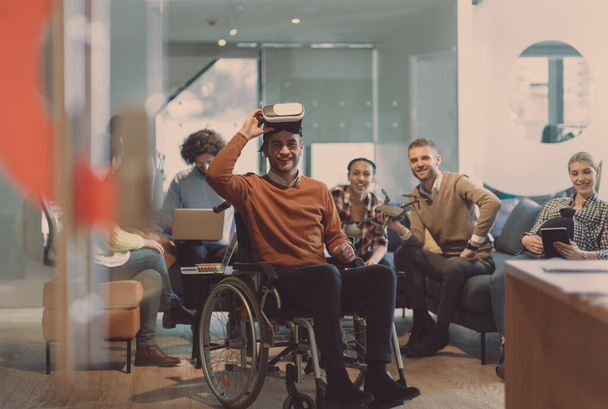 Disabled businessman in a wheelchair at work in modern open space coworking office on team meeting using virtual reality goggles and drone assistance simulation or brainstorming. Effective teamwork of - Valokuva, kuva