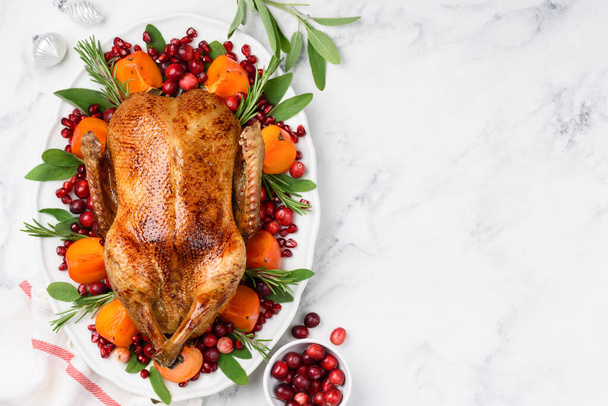 Christmas oven roasted duck on a white plate decorated with cranberries, pomegranate, persimmon, rosemary and sage. Marble background. Copy space. Chrismas food concept.  - Φωτογραφία, εικόνα