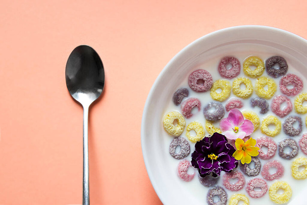sobre un fondo liso melocotón un hermoso desayuno americano de verano: anillos de escamas multicolores con leche y flores violetas de color rosa amarillo y violeta. hay una cuchara al lado del plato  - Foto, imagen