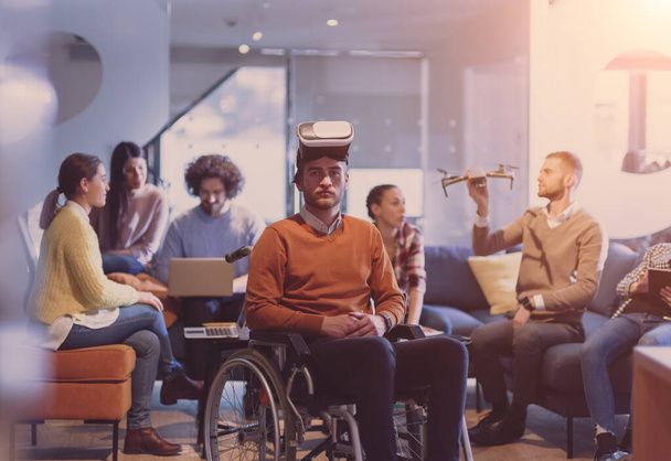 Disabled businessman in a wheelchair at work in modern open space coworking office on team meeting using virtual reality goggles and drone assistance simulation or brainstorming. Effective teamwork of - Photo, Image
