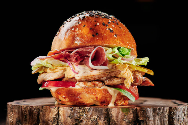 Soczysty burger z kurczakiem i bekonem, serem, pomidorami, sałatą na drewnianym kawałku na czarnym tle. Menu hamburgerów. Miejsce na tekst. - Zdjęcie, obraz
