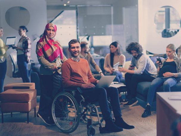 Disabled businessman in a wheelchair at work in modern open space coworking office on team meeting or brainstorming. Muslim businesswoman with hijab is assisting and helping on the project. Effective - Фото, изображение