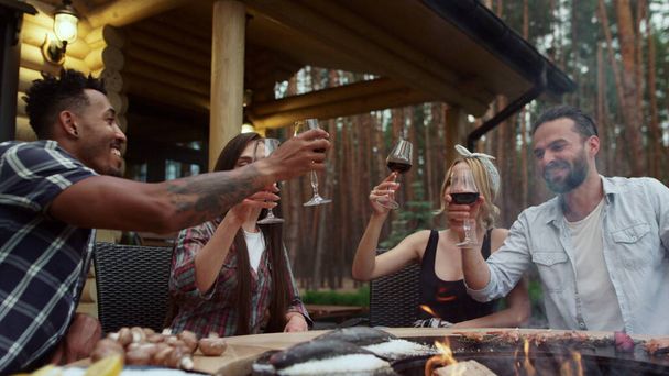Fröhliche Freunde klirren auf einer Grillparty draußen mit Gläsern. Jungs trinken Rotwein - Foto, Bild