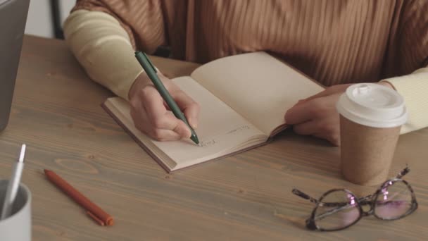 Bloqueado de una persona no reconocible recortada sentada en el escritorio con taza de papel y vasos en la parte superior, escribiendo con pluma en el cuaderno - Metraje, vídeo