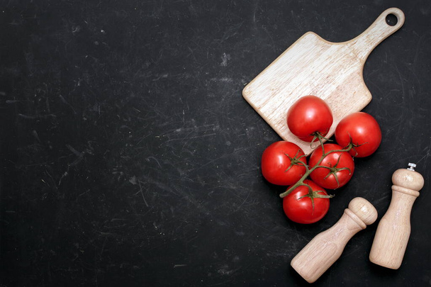 Tomates rojos maduros en rama, papel y molino de sal en el tablero de corte de madera blanca sobre fondo negro. - Foto, Imagen