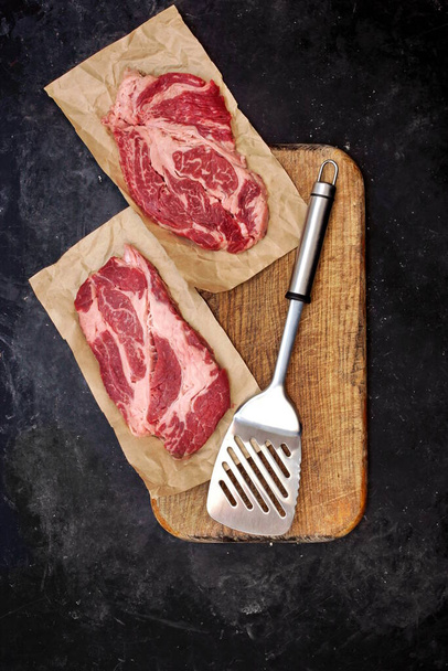 Raw Marbled Loin Beef Steaks and Grill Tools On Wooden Cutting Board. Steaks de boeuf sur spatule prêts pour barbecue ou barbecue, vue aérienne. Steaks de boeuf marbrés Striploin crus sur fond noir, vue du dessus. - Photo, image