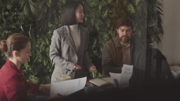 Średnie ujęcie młodej azjatyckiej bizneswoman stojącej na pierwszym planie zielonej ściany rośliny, komunikującej się z kolegami siedzącymi przy stole konferencyjnym, uśmiechającej się, patrzącej na dokumenty - Materiał filmowy, wideo