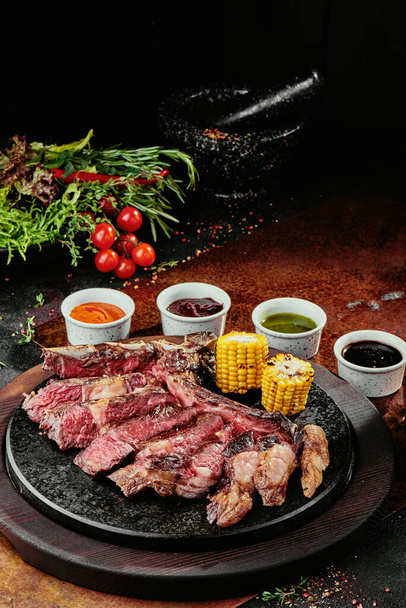 T-Bone Steak serveren met gegrilde groenten en diverse sauzen op een zwarte ronde plaat op een achtergrond van groenten, specerijen en groenten, op een koperplaat. close-up - Foto, afbeelding