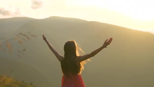 Молодая женщина в красном платье стоит на травянистом лугу в ветреный вечер в осенних горах, поднимая руки, наслаждаясь видом на природу. - Кадры, видео
