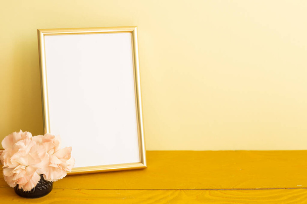 Empty photo frame with carnation flowers on wooden table. yellow background - Photo, Image
