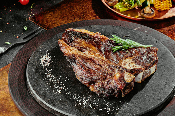 T-Bone steak serving with grilled vegetables on a black round plate on a background of greens, spices and vegetables, on a copper sheet. close up - Foto, afbeelding