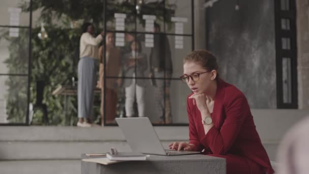 Plan moyen d'une femme blanche professionnelle assise dans un bureau avec un intérieur moderne, tapant sur un ordinateur portable, regardant ses collègues debout sur le fond - Séquence, vidéo