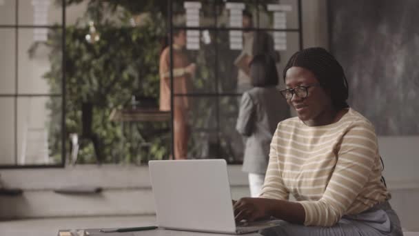 Średnie ujęcie szczęśliwej Afrykanki siedzącej w biurze z nowoczesnym wnętrzem, piszącej na przenośnym komputerze i uśmiechniętej - Materiał filmowy, wideo