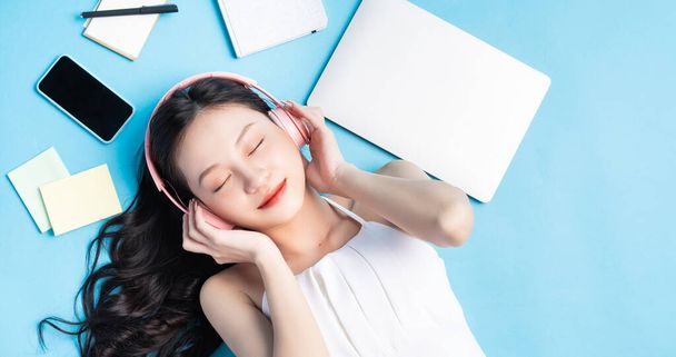 Joven chica asiática acostada sobre fondo azul con ordenador portátil, teléfono inteligente, auriculares y nota alrededor - Foto, imagen
