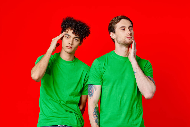 two friends in green t-shirts are standing next to tin with their hands emotions red background - Photo, Image