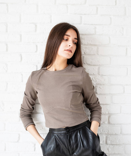 Three quarters length portrait of beautiful smiling brunette woman with long hair wearing brown shirt and black leather shorts, on white brick wall background, hands in pockets - Photo, Image