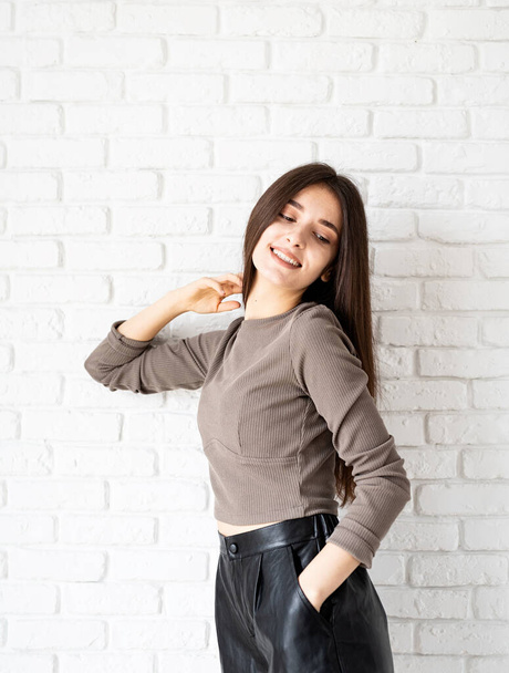 Three quarters length portrait of beautiful smiling brunette woman with long hair wearing brown shirt and black leather shorts, on white brick wall background - Photo, Image