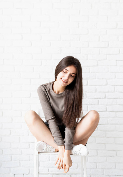 krásná brunetka s dlouhými vlasy v hnědé košili a černé kožené šortky sedí na židli, myšlení a úsměv - Fotografie, Obrázek
