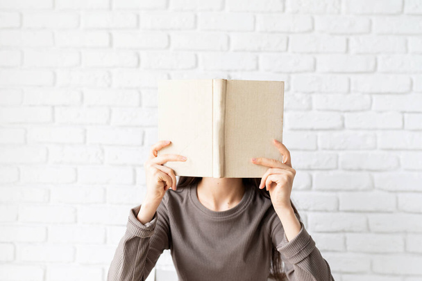 Mujer con camisa marrón sosteniendo un libro abierto delante de su cara - Foto, imagen