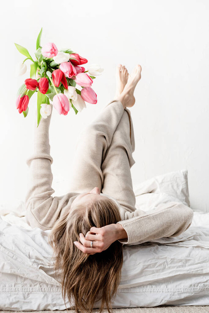 Ευτυχισμένη γυναίκα ξαπλωμένη στο κρεβάτι φορώντας πιτζάμες κρατώντας φωτεινά λουλούδια τουλίπας μπουκέτο σε τεντωμένα χέρια - Φωτογραφία, εικόνα