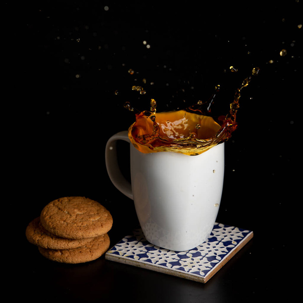 Glass with tea pouring with liquid with splashes and drops of water. Brewing tea concept. Cup pouring with water or tea with splashes on dark wooden background. Cup, teabag and cookies on table. - Fotó, kép