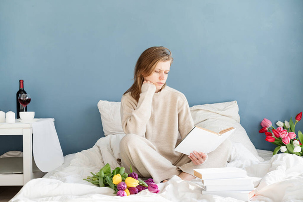 Onnellinen hymyilevä nainen istuu sängyllä yllään pyjama, ilo nauttia kukista ja lukea kirjaa - Valokuva, kuva