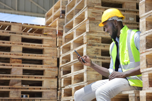 Biztonsági mellényben és keménykalapban dolgozó afro-amerikai munkás mobiltelefonnal ellenőrzi a bejövő rakományt, miközben a raklapfán ül a raktárban. - Fotó, kép