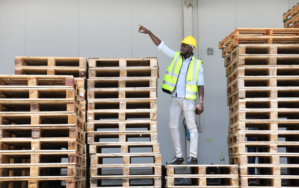 Travailleur afro-américain en gilet de sécurité costume et casque debout sur la pile de bois de palette à l'entrepôt - Photo, image