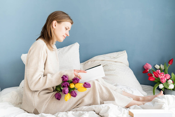 Mutlu, gülümseyen bir kadın pijamalarıyla yatakta oturuyor, çiçeklerin tadını çıkarıyor ve kitap okuyor. - Fotoğraf, Görsel