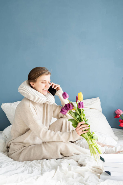 Ευτυχισμένη γυναίκα που κάθεται στο κρεβάτι φορώντας πιτζάμες μιλώντας στο τηλέφωνο, φόντο μπλε τοίχου - Φωτογραφία, εικόνα
