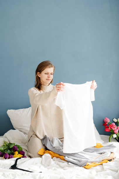 Νεαρή χαμογελαστή γυναίκα που οργανώνει ρούχα που κάθονται στο κρεβάτι στο σπίτι - Φωτογραφία, εικόνα