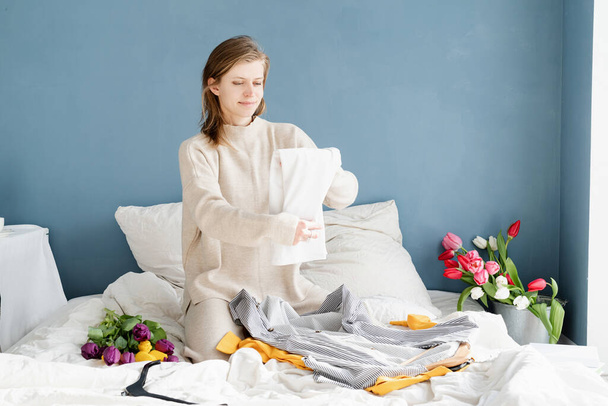 Mladá usměvavá žena organizující oblečení sedící doma na posteli - Fotografie, Obrázek