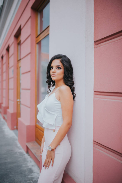 A young brunette female model posing in front of a building with pink walls in an elegant white blouse - Фото, зображення