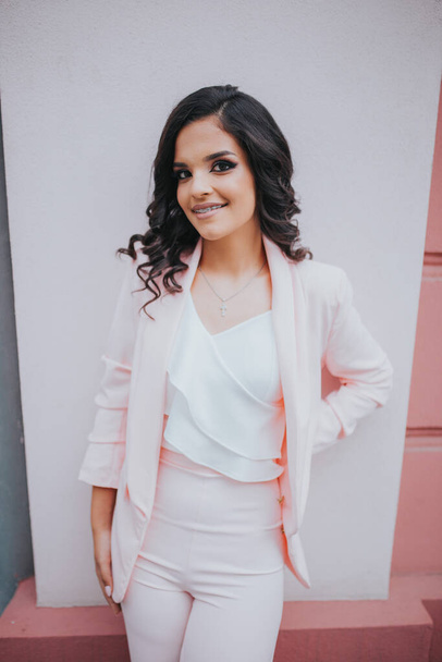A young brunette female model posing in front of a building with pink walls in an elegant white blouse - Photo, Image