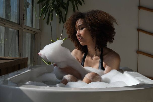 Lachende en ontspannende Afro-Amerikaanse vrouw badend in een bad vol schuim. Geweldige tijd. levensstijl mensen concept - Foto, afbeelding