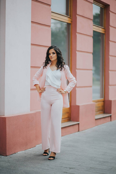 A young brunette female model posing in front of a building with pink walls in an elegant white blouse - Фото, изображение